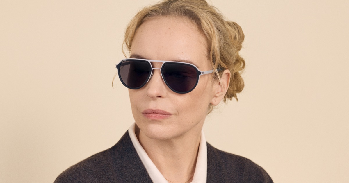 Nina Hoss wearing MYKITA aviator sunglasses with a white collar and dark cardigan against a neutral cream background.