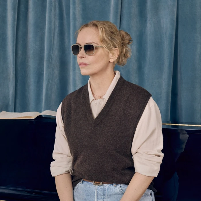 Nina Hoss wearing MYKITA sunglasses, a dark sweater vest over a light shirt, against a blue curtain backdrop.