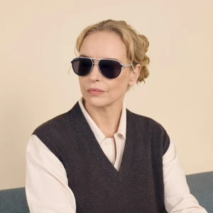 Nina Hoss wearing dark sunglasses, a white shirt, and a dark grey sweater vest against a neutral background for MYKITA Heritage Series.