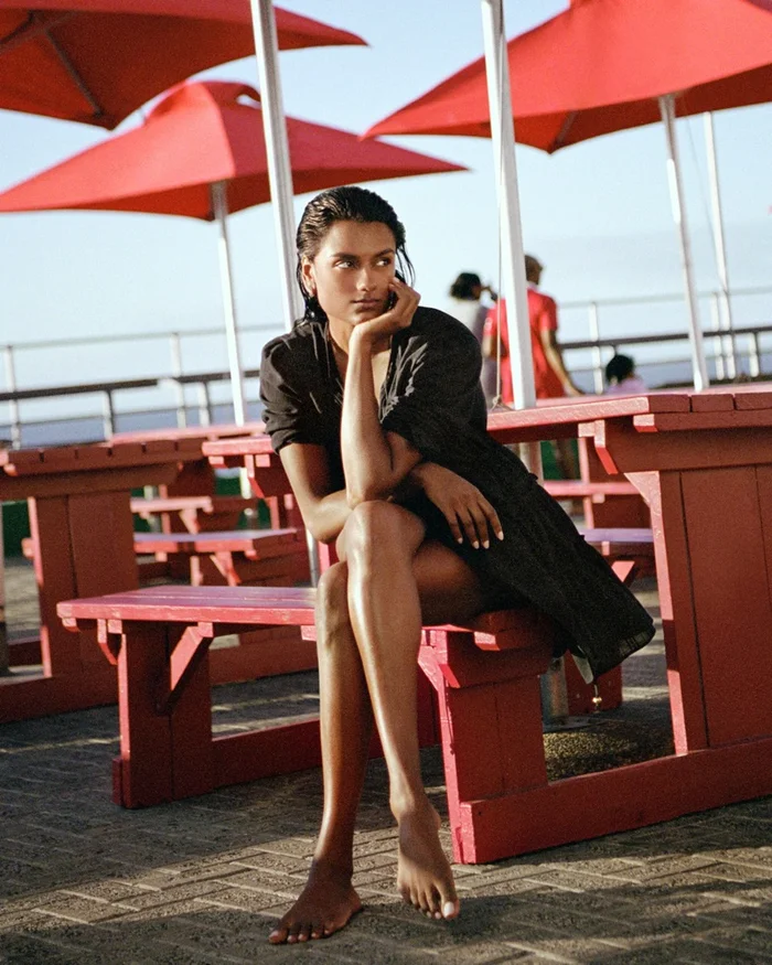 Simone Ashley seated outdoors on a red picnic bench beneath red umbrellas wearing a dark dress