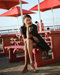 Simone Ashley seated outdoors on a red picnic bench beneath red umbrellas wearing a dark dress