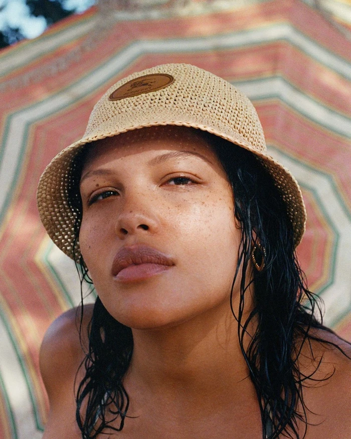 Model wearing a woven straw bucket hat with a leather patch in front of a patterned beach umbrella