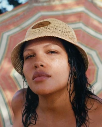 Model wearing a woven straw bucket hat with a leather patch in front of a patterned beach umbrella