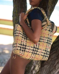 Model standing outdoors wearing a dark t-shirt and holding a woven plaid tote bag against a tree trunk.