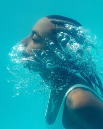 Close-up profile of Simone Ashley underwater with swirling air bubbles and bright turquoise water for Burberry High Summer 2026.