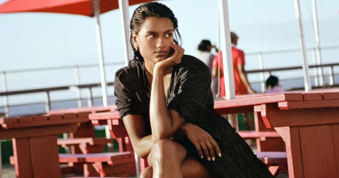 Simone Ashley seated outdoors at a red picnic table wearing a dark top, shot by Ryan McGinley for Burberry High Summer 2026.