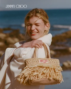 Blonde model smiling while holding a straw handbag against an ocean background for Jimmy Choo Paradise Cove campaign.