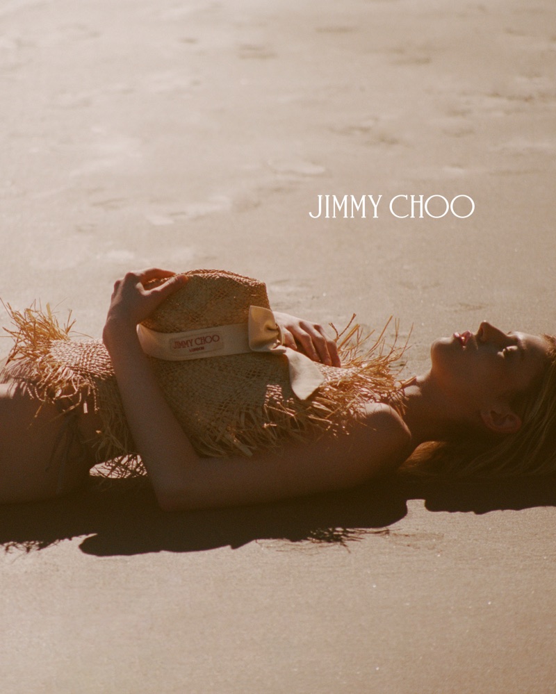 Model lying on a sandy beach holding a straw hat with a ribbon for Jimmy Choo Paradise Cove campaign