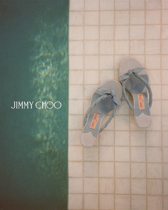 A pair of light blue sandals rests beside a swimming pool on beige tiled ground in a campaign photograph for Jimmy Choo.