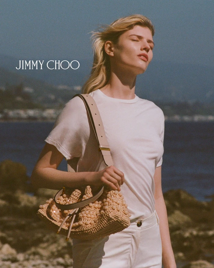 Model wearing a white t-shirt carrying a woven straw bucket bag with beaded details against an ocean background