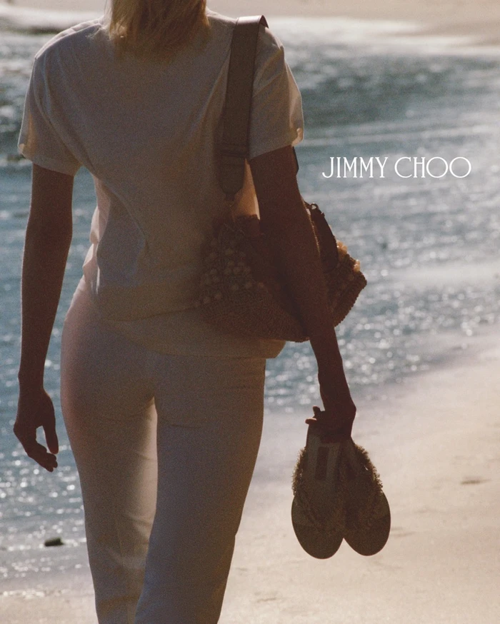 Woman walking on sandy beach holding sandals, wearing white t-shirt and white trousers with shoulder bag for Jimmy Choo Paradise Cove campaign