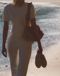 Woman walking on sandy beach holding sandals, wearing white t-shirt and white trousers with shoulder bag for Jimmy Choo Paradise Cove campaign