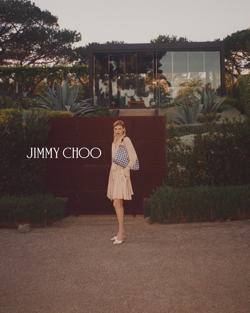 Model standing outdoors holding a blue and white patterned handbag, wearing a light beige dress and white shoes.