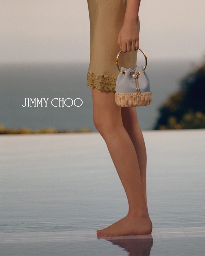 Model standing by a pool holding a light blue bucket bag with a gold ring handle for Jimmy Choo Paradise Cove campaign.