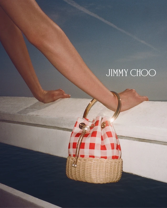 Model leaning arms on a ledge with a red and white checkered bucket bag by Jimmy Choo against an ocean sky background.