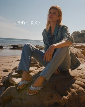 Model wearing a denim shirt and jeans sitting on rocky beach under clear sky for Jimmy Choo Paradise Cove campaign