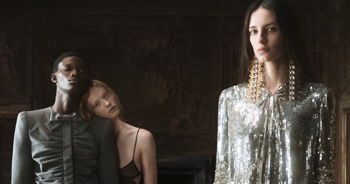 Three models posing in formal Valentino fashion, including a sequined top with chandelier earrings and a tailored grey jacket.