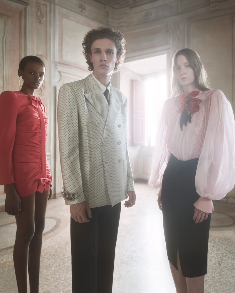 Three models posing in an ornate room wearing a tailored suit jacket, a red ruched dress, and a sheer pink blouse.