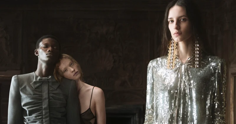 Three models posed in a dimly lit interior, featuring a sequined top and tailored grey jacket.