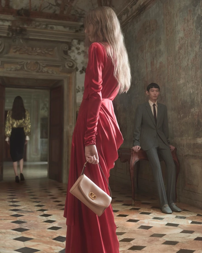 Model in long red silk dress holding light leather handbag in ornate historical interior with tiled floor