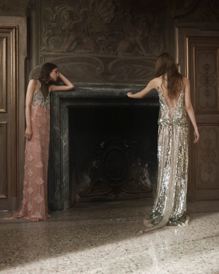 Two models in embellished evening dresses standing by a classical stone fireplace in an ornate interior room