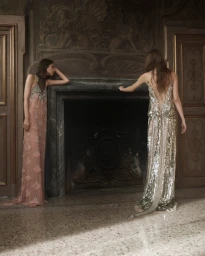 Two models in embellished evening dresses standing by a classical stone fireplace in an ornate interior room