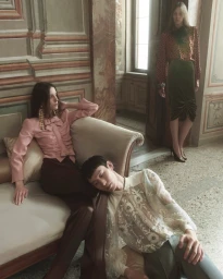 Three models posed in an ornate interior, including a woman on a couch, a man seated on the floor, and a standing woman.