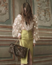 Model in sheer polka dot blouse and yellow skirt holding a brown leather handbag against an ornate wall paneling background.