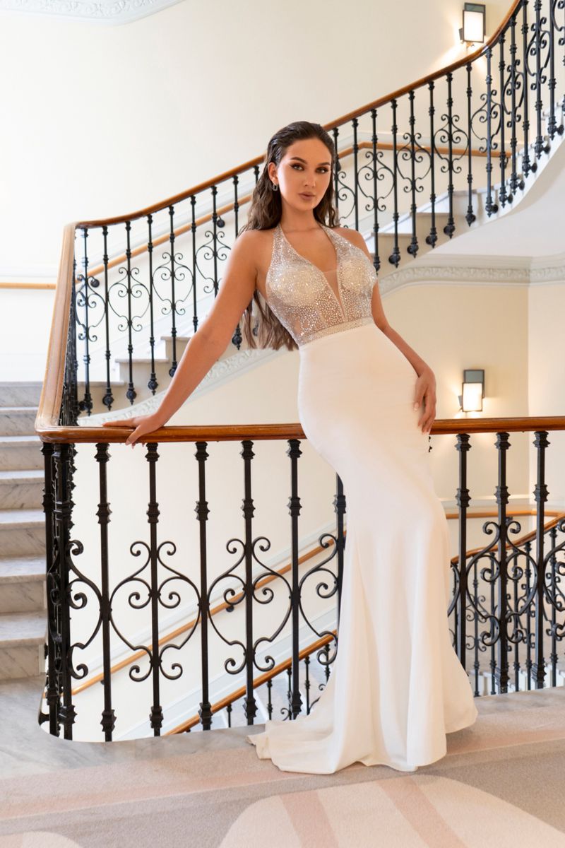 Elegant woman in a white halter-top dress poses on carpeted stairs with a decorative iron railing and a marble staircase, indoor setting.