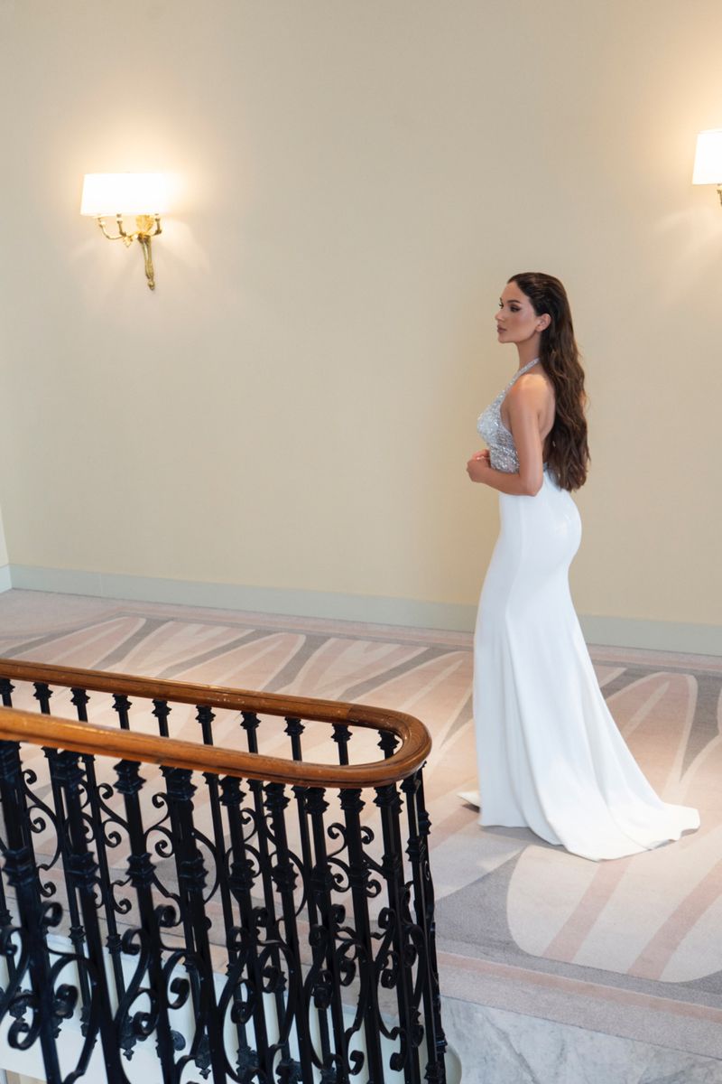 Brunette woman in a white gown poses on stairs indoors, with a black railing and wall sconces, illuminated by soft, natural light.