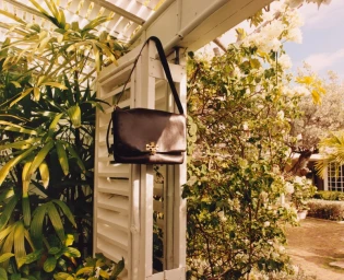 Tory Burch black leather shoulder bag hanging on a white wooden pillar amidst lush tropical plants and blooming white flowers.