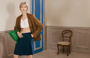 Gigi Hadid wearing a brown blazer, white top, and blue skirt, holding a green leather bag for Miu Miu Leather Goods 2026.