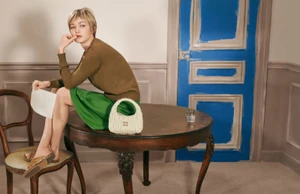 Gigi Hadid seated on a wooden table in an olive sweater and green skirt next to a white handbag for Miu Miu.