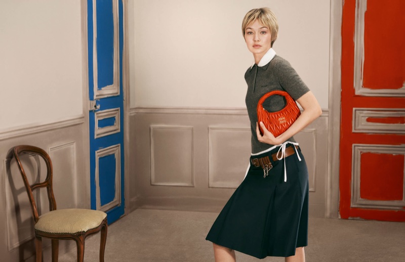 Gigi Hadid wearing a dark polo shirt and skirt holding an orange leather handbag in a room with blue and red doors.