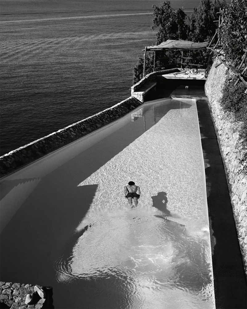 High-angle black and white shot of a man swimming in a long cliffside infinity pool overlooking the ocean.