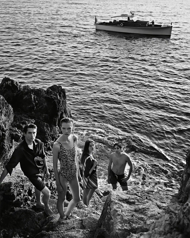 Four models in swimwear and resort attire standing on a rocky shoreline with a boat anchored in the sea in black and white.