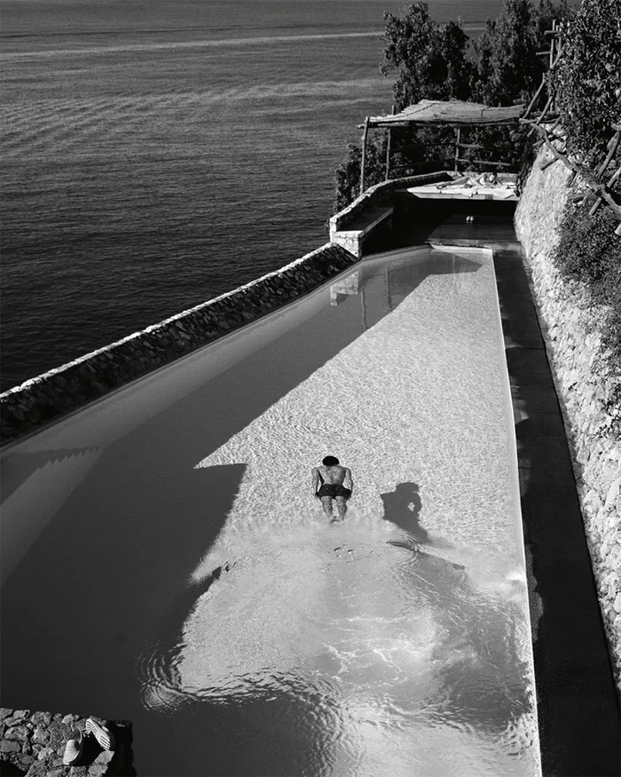 High-angle black and white photograph of a person swimming in a long coastal pool overlooking the sea.