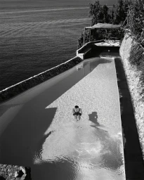 High-angle black and white photograph of a person swimming in a long coastal pool overlooking the sea.