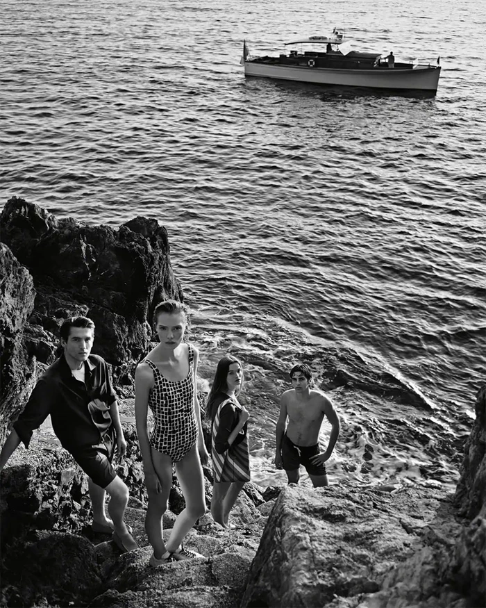 Black and white editorial photograph of four models standing on rocky coastal terrain by the sea with a boat visible in the background.