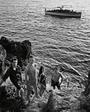 Black and white editorial photograph of four models standing on rocky coastal terrain by the sea with a boat visible in the background.