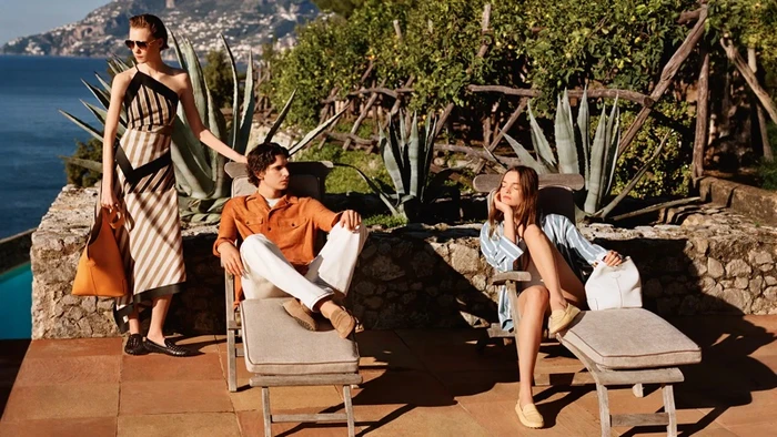 Three models posing outdoors near a coastal terrace, featuring Tod's luxury fashion and accessories.