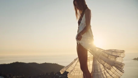 Model standing outdoors against a coastal landscape wearing a sheer white lace dress during sunset.