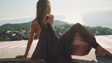 Model wearing a brown halter dress seated on a concrete wall against an ocean and hills background