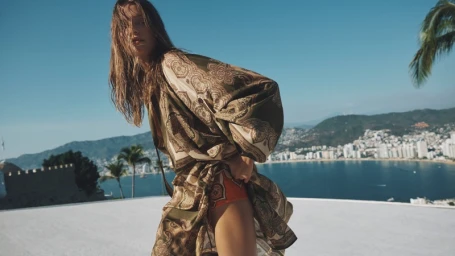 Model in printed green and beige patterned wrap dress posing on a terrace overlooking a coastal city and bay