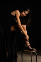 Model Kaia Gerber wearing a black dress and black shoes while posing on a stool against a dark background for Repetto In Bloom.