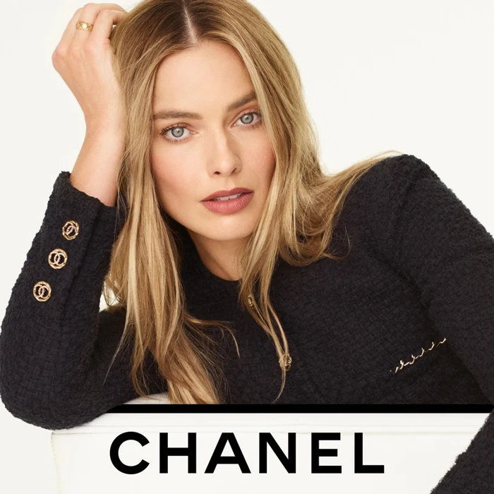 Margot Robbie wearing a black tweed jacket with gold buttons in a campaign for Chanel Rouge Allure Velvet against a white background.