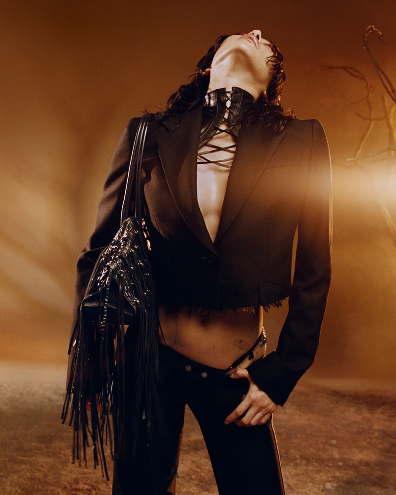 Model with head tilted back wearing a cropped black blazer, lace-up neck corset, and holding a black fringe bag under warm amber light.