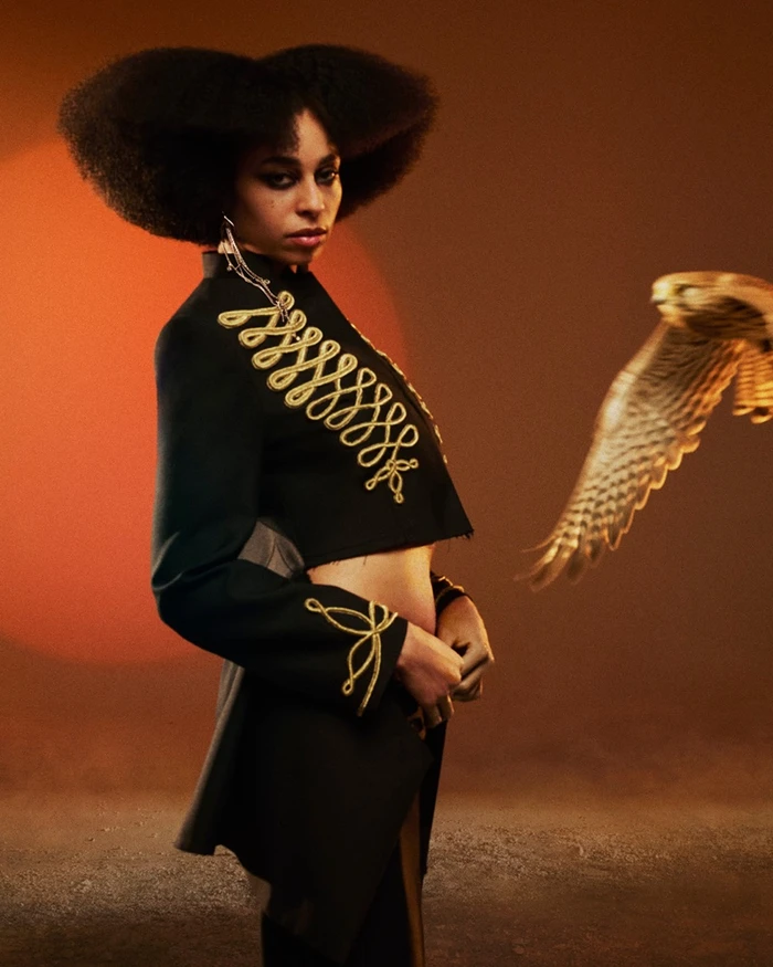 Woman wearing a black military jacket with gold embroidery and a bird flying past in warm orange studio lighting.