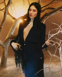 Caroline Polachek in a dark structured blazer and fringed black handbag posing among gnarled tree branches.