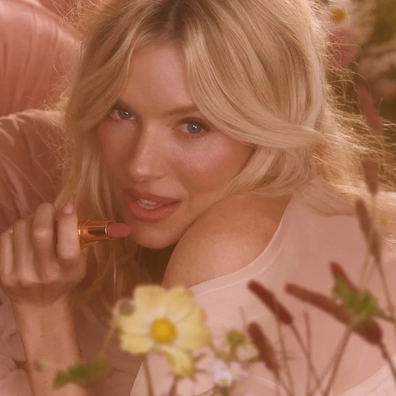 Close-up portrait of Sienna Miller holding a pink lipstick amongst soft floral blooms for the Charlotte Tilbury Pillow Talk in Bloom campaign.